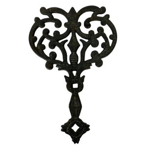 May include: Black cast iron trivet with a decorative heart-shaped design. The trivet has a long, slender handle with a square opening at the top.