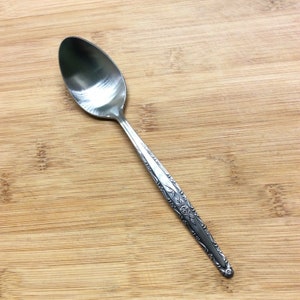 Puede incluir: Una cuchara de plata con un diseño floral en el mango. La cuchara está sobre una superficie de madera.