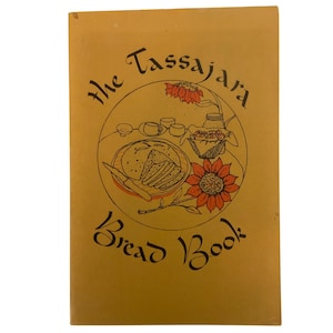 Könnte beinhalten: Ein Vintage-Kochbuch mit dem Titel "The Tassajara Bread Book" mit einem beigefarbenen Einband. Das Cover zeigt eine kreisförmige Illustration mit Brot, Blumen und Text in Schwarz und Orange. Der Titel des Buches ist in einer stilisierten Schrift geschrieben.