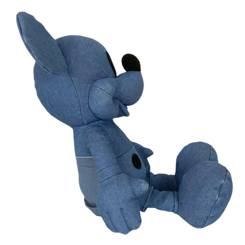 May include: A blue denim plush toy featuring a cartoon character with large pointed ears, a rounded snout, and a black nose. The figure is in a seated position with visible stitched seams, a black circular belly detail, and large feet.