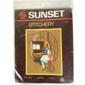 Op de afbeelding: Een vintage Sunset Stitchery naaldviltstickset met een meisje dat piano speelt. De set is verpakt in een bruine plastic zak met de titel "Piano Lesson" en het nummer 2512. De set past in een lijst van 28 cm x 36 cm.
