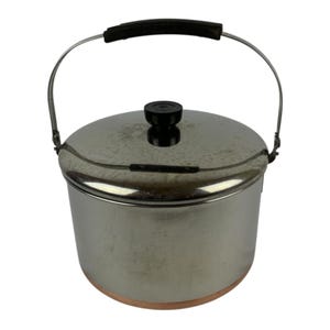 Puede incluir: Una olla de acero inoxidable con base de cobre, que incluye una tapa a juego con un pomo negro. La olla cuenta con un asa giratoria de metal plateado con agarre negro. La superficie exterior es metálica y reflectante.