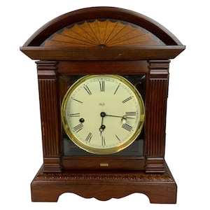 May include: A dark brown wooden mantel clock featuring a cream-colored circular face with black Roman numerals and the brand name Seth Thomas. The clock has a gold-toned bezel, fluted side columns, and a decorative fan-shaped inlay at the top.