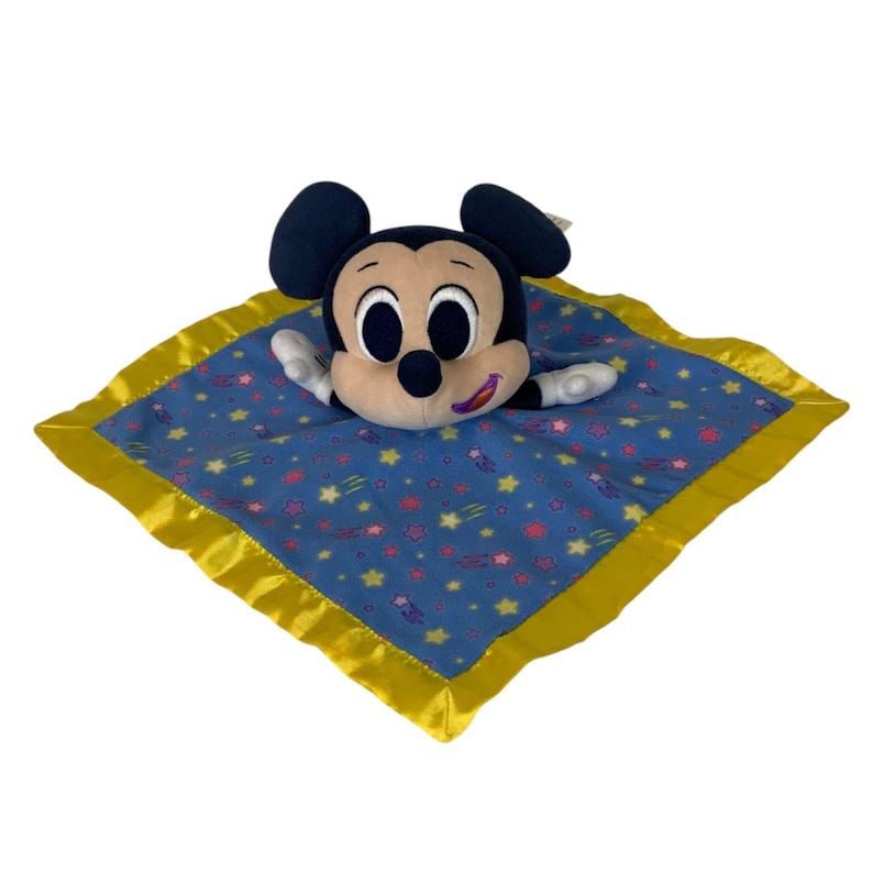May include: A plush security blanket featuring a Mickey Mouse head with large black ears and a tan face. The square blanket is blue with a pattern of small yellow and pink stars and has a shiny yellow satin border trim around the edges.