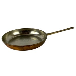 May include: A round frying pan with a metallic copper exterior and a silver-toned interior lining. The pan features a long, slender handle attached to the side with two visible rivets. The surface shows signs of wear and natural metal oxidation.