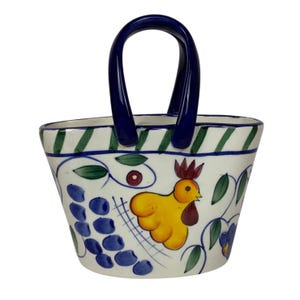 May include: A ceramic basket with a dark blue handle and rim. The white body is decorated with a yellow cockerel, blue grapes, and green leaves. The rim has green and blue diagonal stripes.