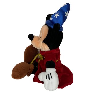 Peut inclure: Un Mickey Mouse en peluche habill&eacute; en sorcier. Il porte un chapeau bleu avec des &eacute;toiles blanches et une robe rouge. Il a un sac brun dans sa main.