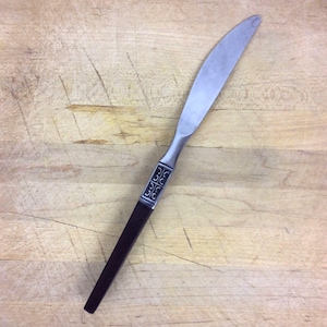 May include: A silver butter knife with a black handle and a decorative pattern. The knife is on a wooden surface.