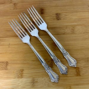 Puede incluir: Tres tenedores plateados con detalles florales ornamentados en los mangos. Las púas son largas y delgadas. Los tenedores están dispuestos diagonalmente sobre una superficie de madera, mostrando su elegante diseño.