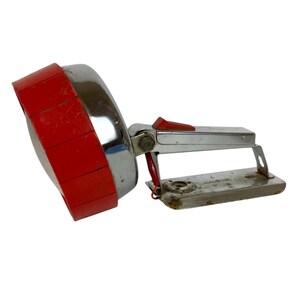 Puede incluir: Un foco de mano vintage que presenta una gran carcasa reflectora cromada circular con un borde de plástico rojo brillante. El dispositivo incluye un mango metálico con un interruptor de gatillo rojo y una placa base metálica rectangular.