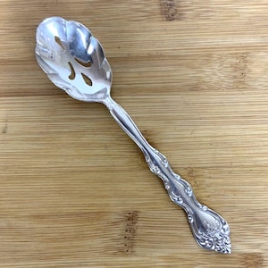 Può includere: Un cucchiaio argentato con fessure e un manico decorato. Il cucchiaio ha un design decorativo con un bordo smerlato e una serie di fori nella ciotola.