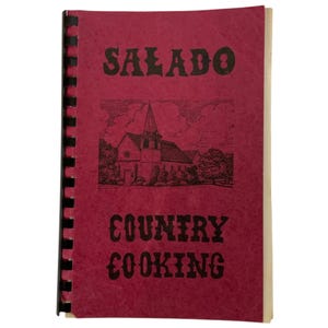 May include: A red spiral-bound cookbook titled "SALADO COUNTRY COOKING." The cover features a black illustration of a church with a steeple, trees, and clouds. The text is in a bold, vintage font.