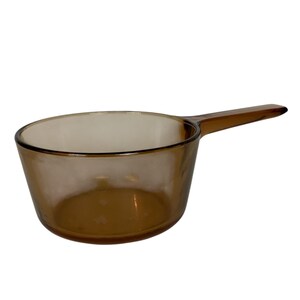 May include: A translucent amber-coloured glass saucepan featuring a round bowl and a single elongated handle extending from the side. The vessel has a smooth, polished finish and a consistent brown tint throughout the entire glass structure.