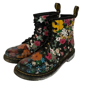 Könnte beinhalten: Paar schwarze Leder-Kampfstiefel mit farbenfrohem Blumenmuster. Die Stiefel haben schwarze Schnürsenkel, gelbe Nähte und eine schwarze Sohle. Das Blumenmuster umfasst rosa, gelbe, orange und blaue Blumen.