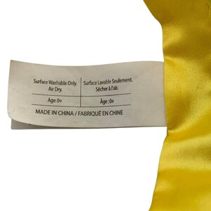 May include: A white rectangular fabric care label attached to a bright yellow satin material. The label features black text reading Surface Washable Only. Air Dry. Age: 0+ and MADE IN CHINA, alongside French translations for the same instructions.