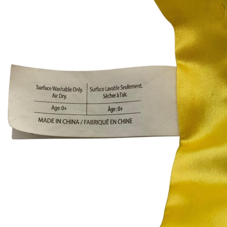 May include: A white rectangular fabric care label attached to a bright yellow satin material. The label features black text reading Surface Washable Only. Air Dry. Age: 0+ and MADE IN CHINA, alongside French translations for the same instructions.