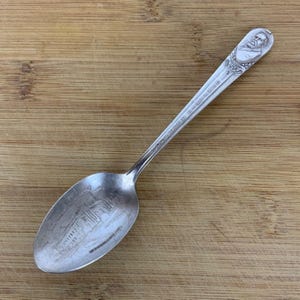May include: Silver souvenir spoon with a depiction of a building and a man's face on the handle. The spoon has the text "The Home of the Brave" on the handle.