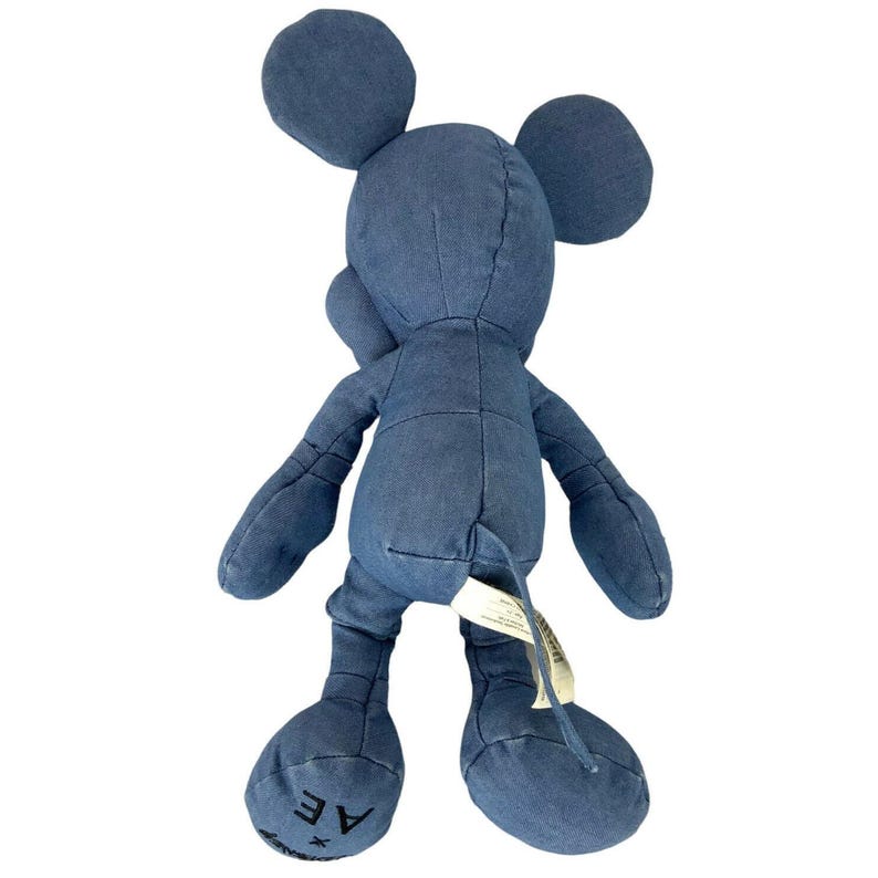 May include: A blue denim plush toy shaped like a mouse with large round ears, long arms, and legs. The fabric has visible seams throughout the body. The left foot features the black text AE, and a white fabric tag is attached near the tail area.