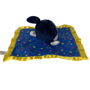 May include: A plush security blanket featuring a dark blue Mickey Mouse head with rounded ears attached to a square blue fabric base. The blanket is decorated with a pattern of yellow stars and pink shapes, finished with a bright yellow satin border.