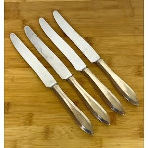 May include: Four silver butter knives with a shiny blade and a rounded handle are arranged diagonally on a wooden cutting board. The knives have a classic design, suitable for formal dining or everyday use.