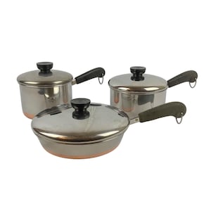 Può includere: Un set di tre pentole in acciaio inossidabile con basi color rame, composto da due casseruole profonde e una padella bassa. Ogni pezzo presenta un coperchio in metallo lucido con pomello nero e un manico nero con un anello per appenderlo.