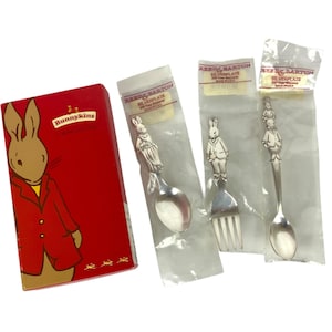 Może przedstawiać: Czerwone pudełko Bunnykins z ilustracją królika oraz trzy posrebrzane sztućce marki Reed & Barton w indywidualnych plastikowych opakowaniach. Zestaw zawiera dwie łyżki i jeden widelec, każdy z figurką królika na uchwycie.