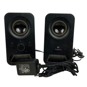 Op de afbeelding: Zwarte Logitech computerspeakers met een voedingsadapter. De speakers hebben een ronde speaker en een volumeknop. Het Logitech-logo is zichtbaar op één speaker.