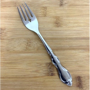 May include: A silver fork with an ornate handle. The fork is on a wooden surface.