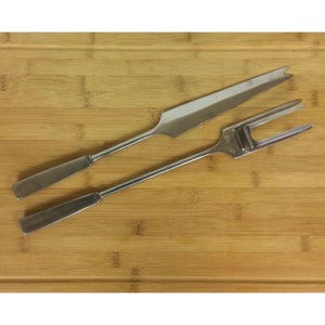 May include: A set of two stainless steel cheese tools. The first tool is a knife with a pointed tip and a serrated edge. The second tool is a fork with four tines and a small hole in the center. The tools are on a wooden cutting board.