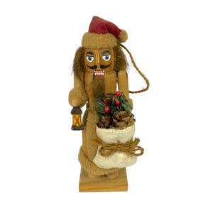 Puede incluir: Una figura de cascanueces de madera con un gorro rojo y ribete de piel sintética beige, cabello castaño, bigote negro y ojos azules. La figura sostiene una pequeña linterna y un saco de tela color crema lleno de piñas y bayas rojas.