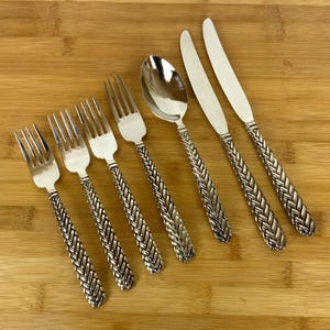 May include: A set of silver-colored flatware, including forks, spoons, and knives, arranged on a wooden surface. The handles feature a woven design. The utensils are likely made of stainless steel or a similar metal.