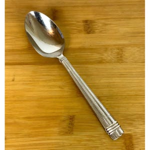 May include: A polished silver-tone metal spoon featuring a smooth, rounded bowl and a long, slender handle. The handle is decorated with a textured, repeating herringbone pattern and finished with a horizontal banded detail at the very end.