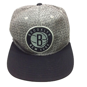 May include: A grey and black baseball cap with a flat peak. The cap has a knitted pattern on the crown and a circular patch with the text "BROOKLYN NEW YORK" around a stylized "B" basketball logo.