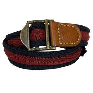 May include: A woven belt with alternating black and burgundy stripes. The belt features a brass buckle and a brown leather tab. The belt is rolled up, showcasing its design and construction. A stylish accessory for any wardrobe.