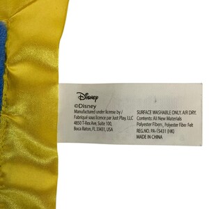 May include: A white fabric product tag attached to a yellow satin-trimmed blue plush item. The tag features black text including Disney, Manufactured under license by Just Play, LLC, 4850 T-Rex Ave, Boca Raton, FL 33431, USA, Surface washable only, and Made in China.