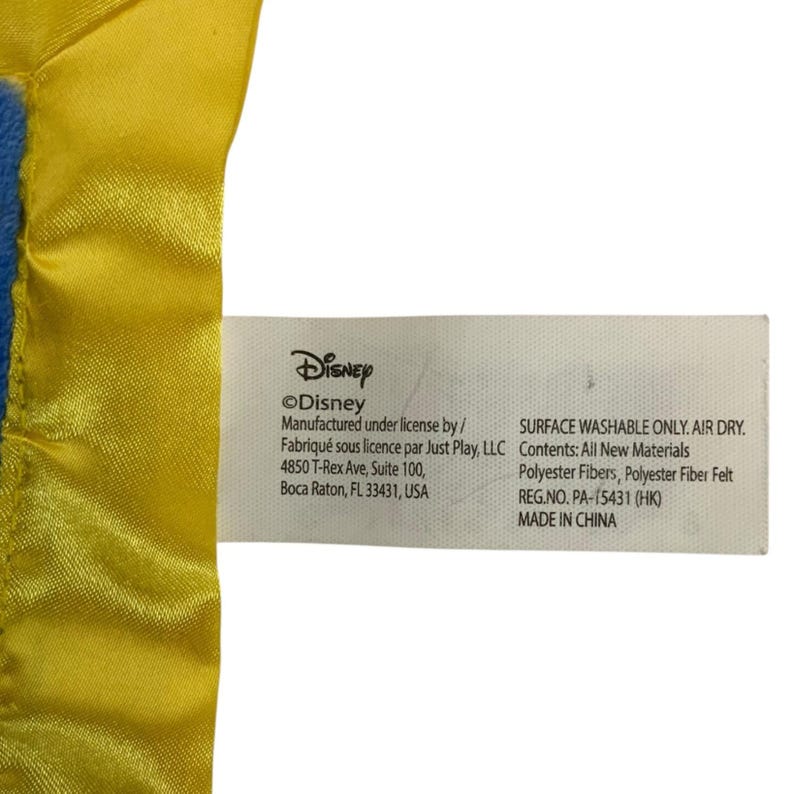 May include: A white fabric product tag attached to a yellow satin-trimmed blue plush item. The tag features black text including Disney, Manufactured under license by Just Play, LLC, 4850 T-Rex Ave, Boca Raton, FL 33431, USA, Surface washable only, and Made in China.