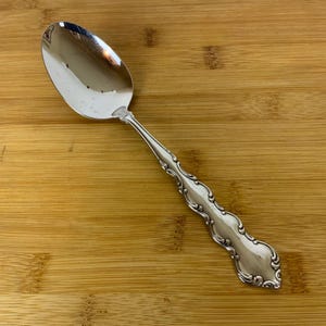 Puede incluir: Una cuchara de plata con un mango adornado. La cuchara tiene un cuenco redondeado y un mango largo y delgado con un diseño decorativo.