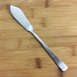 May include: A stainless steel butter knife with a rounded blade and a long, rectangular handle. The knife is set against a wooden background, showcasing its polished metal finish. This utensil is designed for spreading butter or other soft spreads.