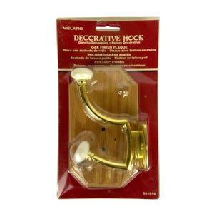 May include: A decorative hook with a polished brass finish and white ceramic knobs mounted on an oak finish plaque. The product is sealed in original retail packaging with the text DECORATIVE HOOK, OAK FINISH PLAQUE, and POLISHED BRASS FINISH.
