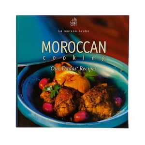 May include: A square cookbook titled Moroccan Cooking: Our Dadas' Recipes by La Maison Arabe. The cover features a vibrant turquoise tagine filled with a savory dish of cooked meat, bright red olives, and yellow fruit, topped with green herbs.