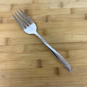 May include: A silver fork with a simple design and a star-shaped pattern on the handle. The fork is lying on a wooden surface.
