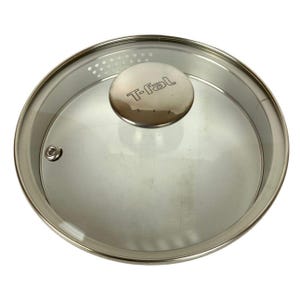 May include: A round T-fal pot lid made of clear tempered glass with a stainless steel rim. The lid features a central metallic knob with the T-fal logo embossed on top, a small steam vent hole, and a section of integrated straining holes.