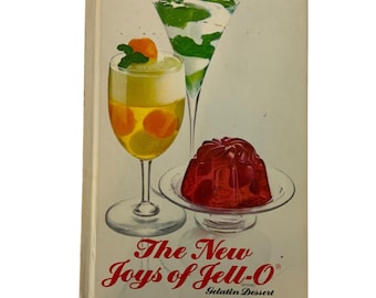 Op de afbeelding: Vintage receptenboek getiteld "The New Joys of Jell-O" met illustraties van gelatinedesserts. De omslag toont drie glazen gevuld met gelaagde gelatine en fruit, en een rode gelatinevorm op een klein bord.