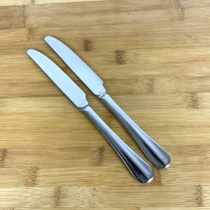 May include: Two stainless steel butter knives with serrated blades and rounded handles are displayed on a wooden cutting board. The knives are angled towards the upper right corner, showcasing their polished finish and simple design.