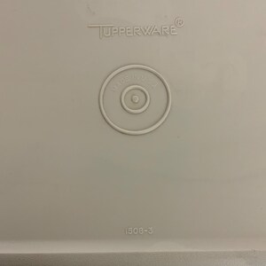 Pode incluir: Uma tampa de recipiente Tupperware cinza claro em close-up. A tampa apresenta o logotipo da Tupperware, um selo circular "Made in U.S.A." e o n&uacute;mero "1508-3".