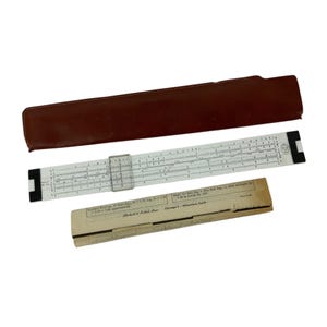 May include: A white Pickett & Eckel slide rule featuring a clear plastic cursor, black end caps, and various numerical scales. It is presented with a dark brown leather-style protective case and a tan paper instruction sleeve containing printed text.