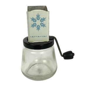 Pode incluir: Picador de cebola de vidro vintage com tampa e cabo pretos. A parte superior de metal branco apresenta um design floral azul. O picador tem uma base de jarro de vidro transparente e uma lâmina de metal.