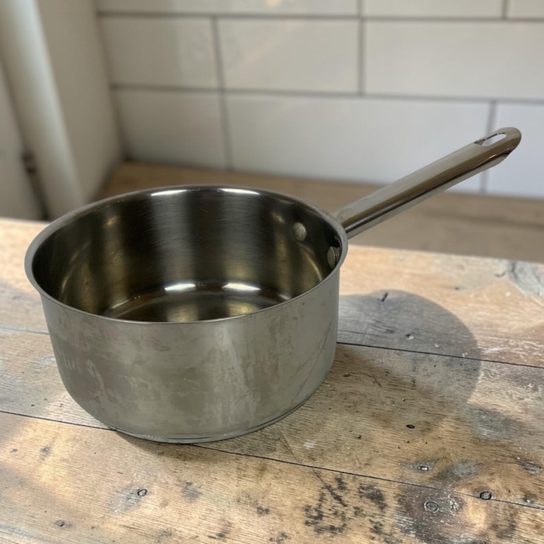 Op de afbeelding: Een roestvrijstalen steelpan met een lange handgreep. De pot is rond met een glanzend, reflecterend oppervlak. De handgreep is aan de zijkant van de pot bevestigd en heeft een lus aan het uiteinde om op te hangen. De pot staat op een houten oppervlak.