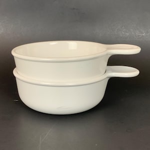 May include: Two white ceramic soup bowls stacked on top of each other. Each bowl has a rounded shape with a handle on one side. The bowls have a smooth, glossy finish and a simple, classic design. The bowls are suitable for serving soup or other dishes.