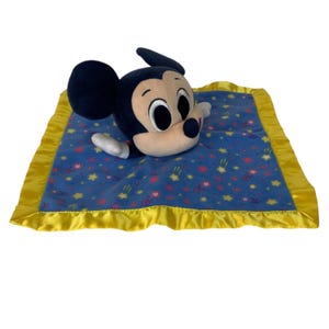 May include: A plush security blanket featuring a Mickey Mouse head and arms attached to a square blue fabric base. The blanket has a yellow satin border and a pattern of small yellow stars and pink flowers printed across the blue fabric surface.
